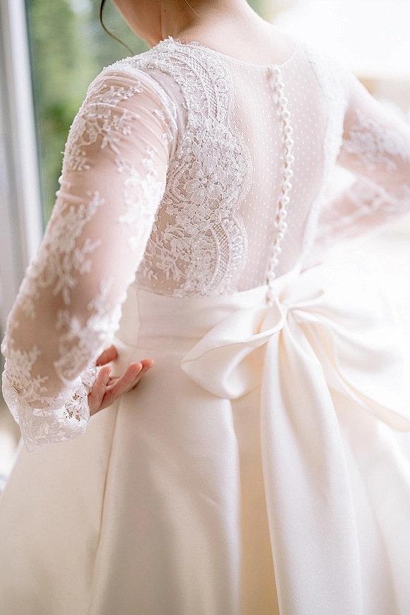 Wedding dress details with lace wedding dress illusion bodice, sheer long sleeves, and covered buttons, lit by a window with blurred greenery