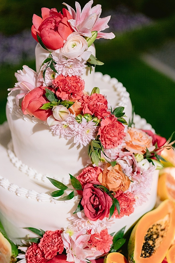 Wedding cake with floral wedding cake topper, scalloped buttercream piping, fresh flowers and papaya slices against blurred garden greenery