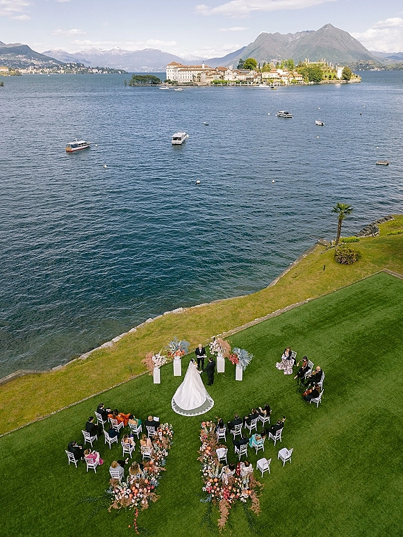 Outdoor wedding ceremony by a lakeside lawn with floral-lined aisle, white chairs, bride’s long train, guests and musicians near the altar