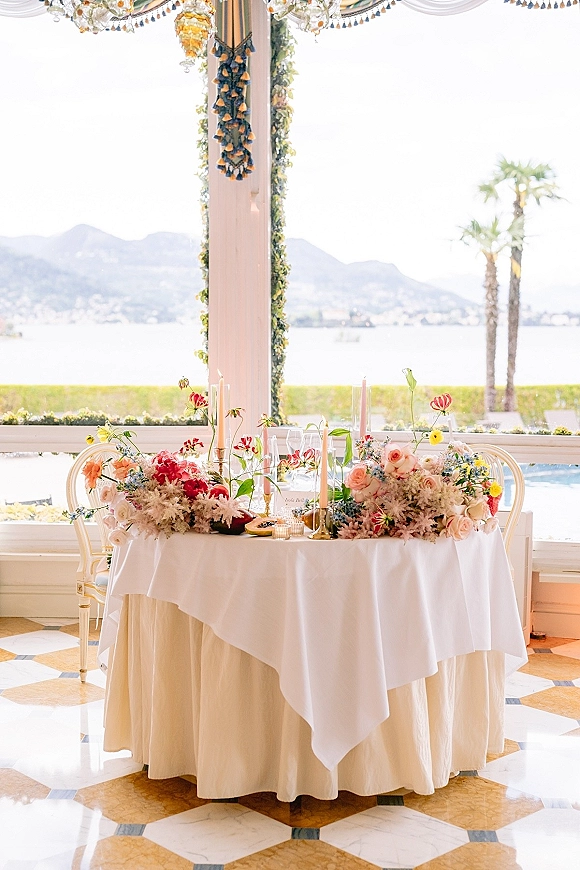 Reception sweetheart table with wedding sweetheart table decor, roses and greenery garland, taper candles, and lake-and-mountain view windows behind