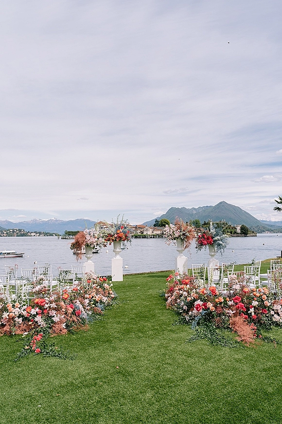 Ceremony setup with floral urn arrangements and white pedestal plinths lining an aisle on a lakeside lawn with mountain views in back