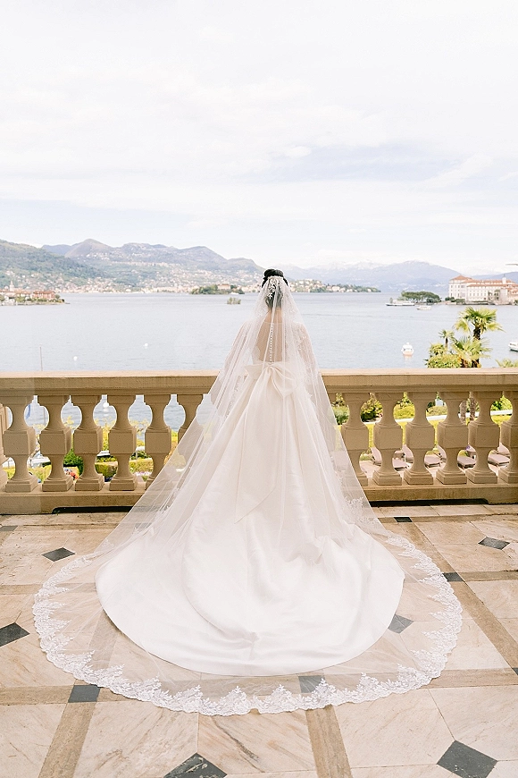 Bridal portrait of a bride from behind in a cathedral veil, lace train flowing on a stone balcony overlooking a lake and mountains