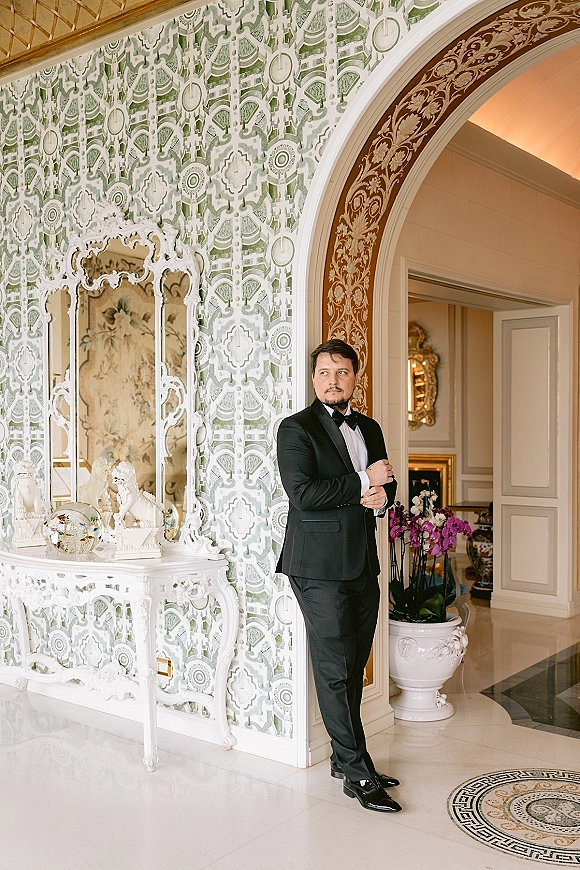 Groom portrait in a classic black tuxedo and bow tie, leaning by an arched doorway in a patterned hallway with ornate mirror decor