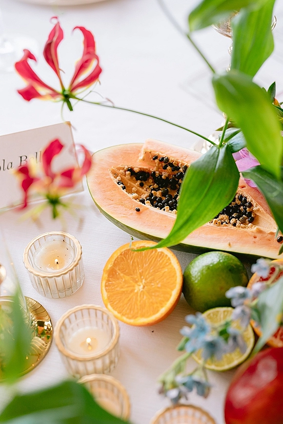 Wedding tablescape with papaya and orange-lime fruit centerpiece, tropical flowers, greenery, gold flatware, and votive candles on white linen