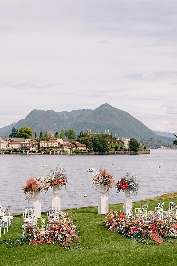 Outdoor ceremony setup with lakeside wedding ceremony chairs, white plinths and pedestal urn florals on a lawn by lake and mountains