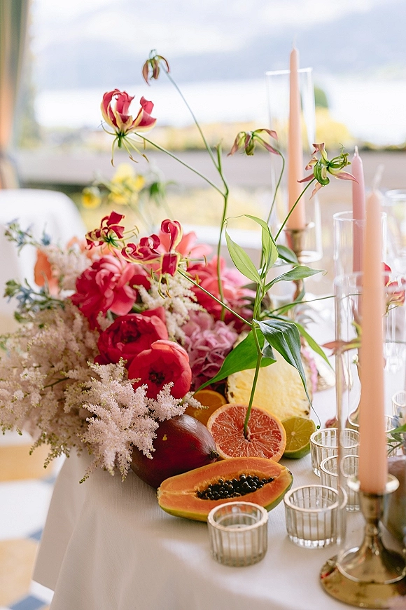 Reception centerpiece with a wedding floral centerpiece of pink and coral blooms, tropical fruit, and taper candles on a terrace table at sunset
