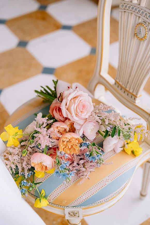 Wedding bouquet in pastel tones with roses, peonies, ranunculus, and greenery resting on a vintage chair over geometric tile floor
