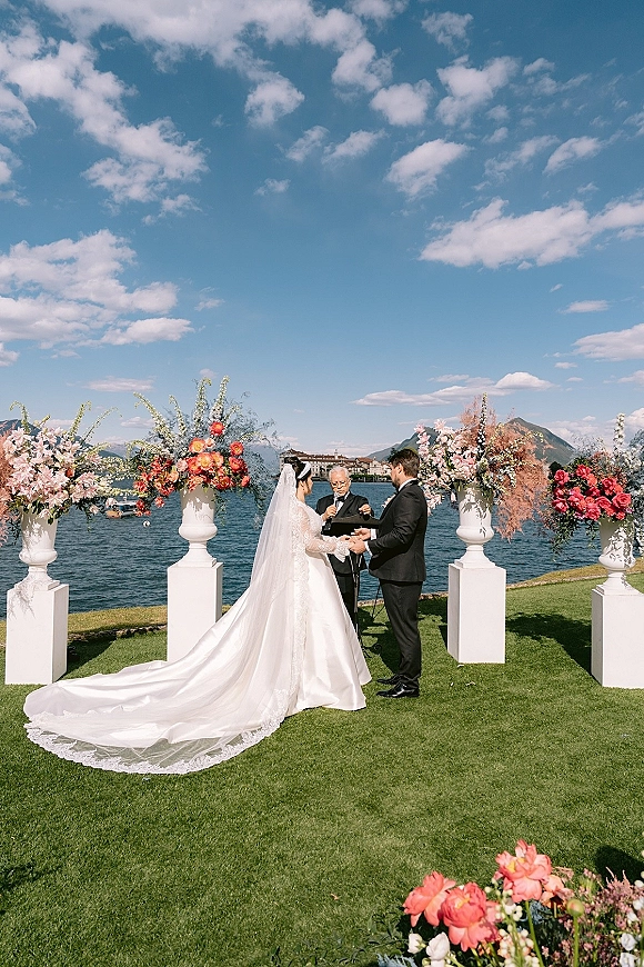 Wedding vows during an outdoor wedding ceremony as bride and groom hold hands, her cathedral veil flowing by floral urns lakeside with mountains