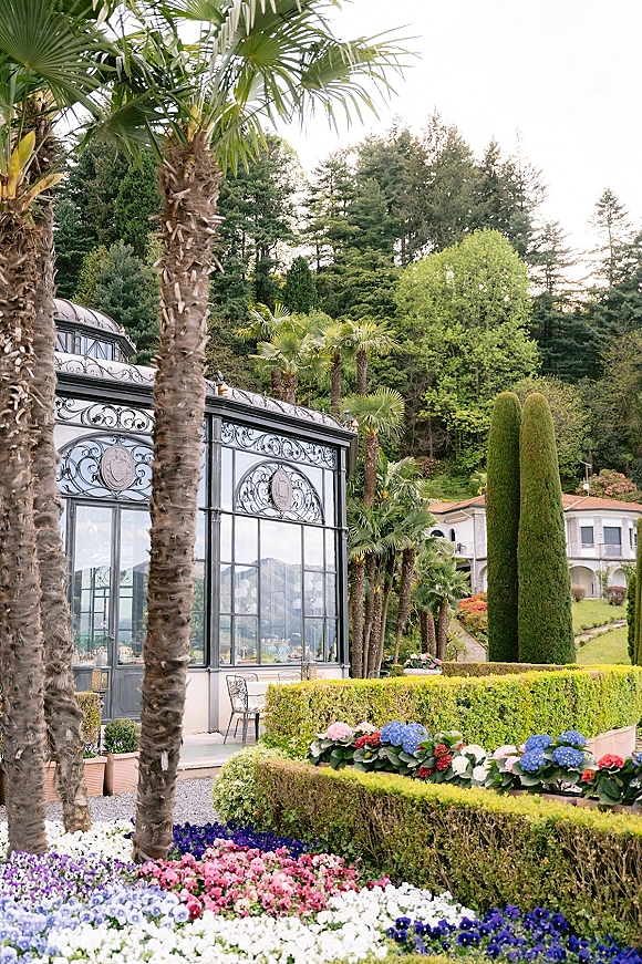 Wedding venue exterior with a glass conservatory, wrought iron details, and palm trees over manicured hedges and garden chairs on a lawn