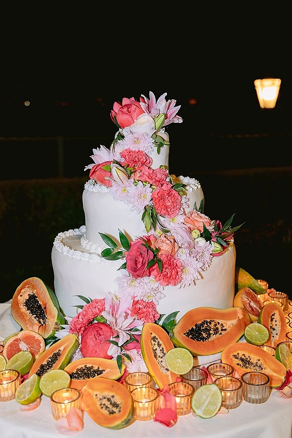 Wedding cake with a floral cascade of pink and coral blooms, three-tier white buttercream, papaya and lime on a candlelit table at night