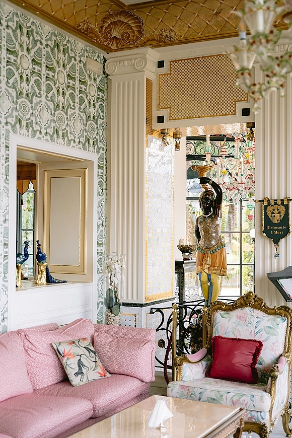 Wedding venue interior lounge with a pink sofa and floral armchair beneath a grand chandelier, set against green patterned wallpaper and gold ceiling detail
