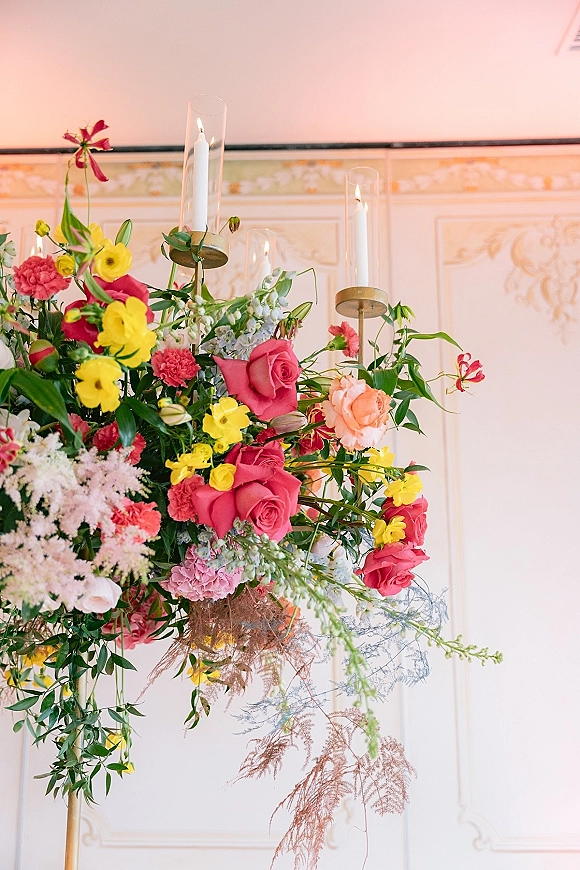 Wedding floral centerpiece with colorful flowers, roses and carnations, plus taper candles in glass hurricanes and gold holders against ornate wall