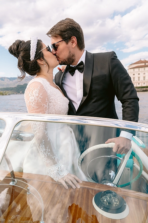 Wedding kiss portrait of bride and groom kissing on a vintage speedboat, bride in lace sleeves with pearl headband, lake and mountains behind