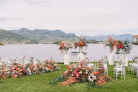 Outdoor ceremony setup with white chairs and a floral ceremony aisle of garden roses and greenery by a lakeside with mountains under clouds