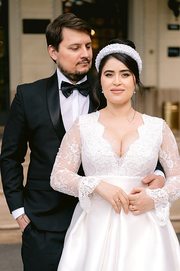 Couple portrait of bride in lace-sleeve wedding dress and pearl headband holding hands with groom in tuxedo at a doorway steps backdrop