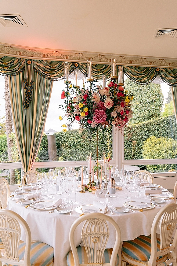 Reception tablescape with a round wedding table setting, tall floral centerpiece on a gold stand, candles and place cards by large windows