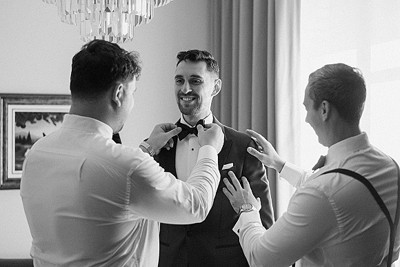 Groom getting ready as groomsmen helping groom adjust his bow tie, wearing a tuxedo in soft window light inside a hotel room