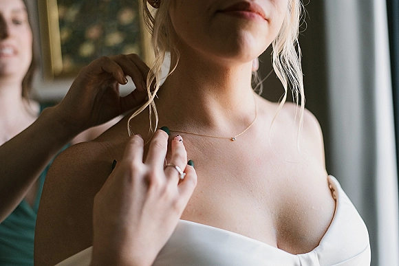 Bridal necklace as a bridesmaid helps fasten the clasp on a bride in a strapless dress, engagement ring visible in window light
