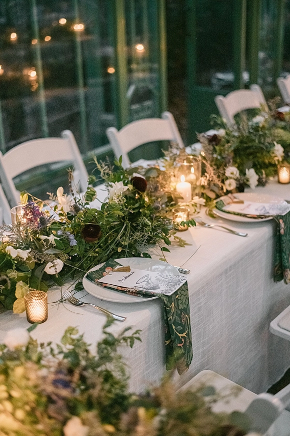 Reception tablescape with a greenery table runner, mixed florals, and candlelight on white linens in a glass greenhouse with bokeh lights