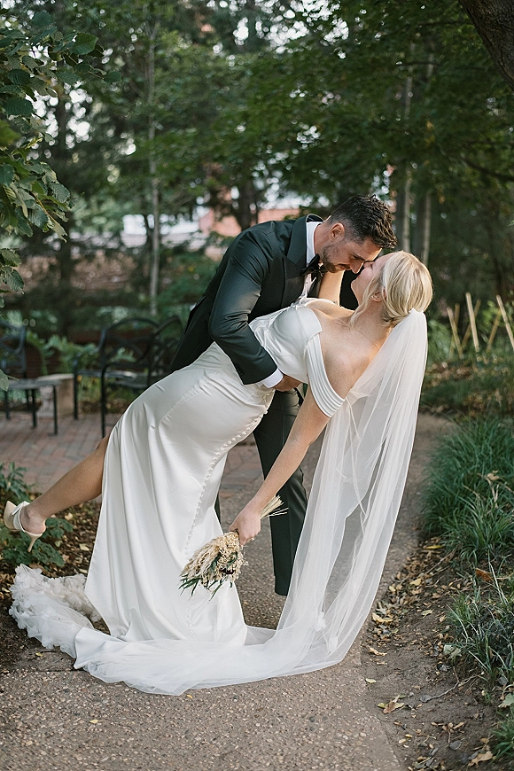 Wedding couple portrait in a wedding dip pose as the groom kisses the bride, her long veil and bouquet flowing along a garden path