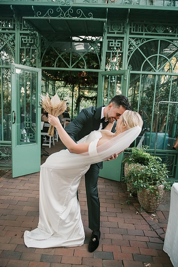 Wedding kiss portrait of bride and groom in a dip kiss, veil flowing as they embrace outside a greenhouse with glass doors and string lights