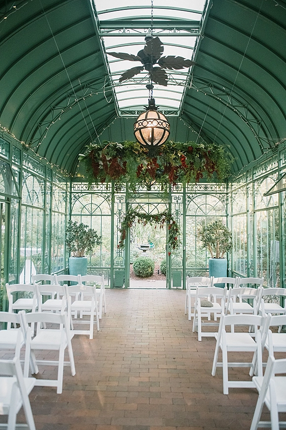 Ceremony setup for a greenhouse wedding ceremony with white folding chairs lining a brick aisle under hanging greenery and string lights in a glass conservatory