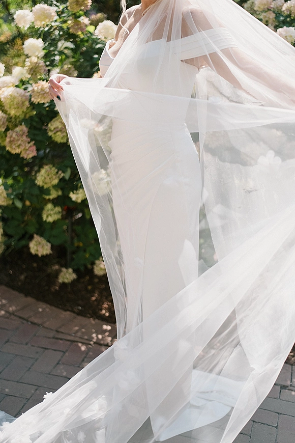 Bridal gown with off the shoulder wedding dress and long veil train, satin fitted silhouette on a sunlit brick walkway by hydrangeas
