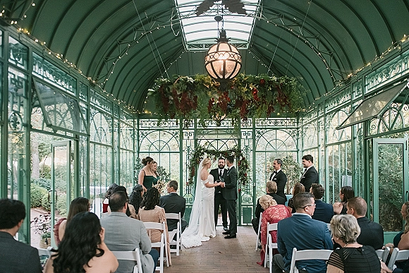 Ceremony moment at a greenhouse wedding ceremony with couple holding hands under a greenery arch, hanging florals and string lights in glass conservatory