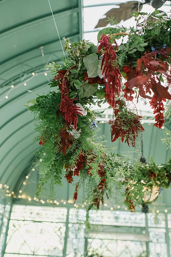 Hanging floral installation with greenery ceiling installation, red blooms and cascading vines on a floral hoop beneath greenhouse windows and string lights