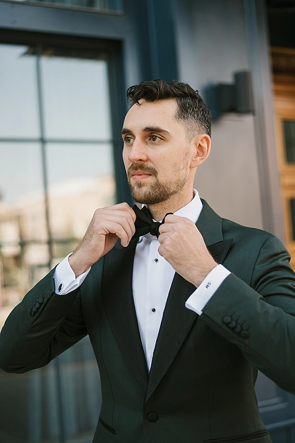 Groom portrait of a groom adjusting bow tie in a black tuxedo with cufflinks beside a building exterior window and wall