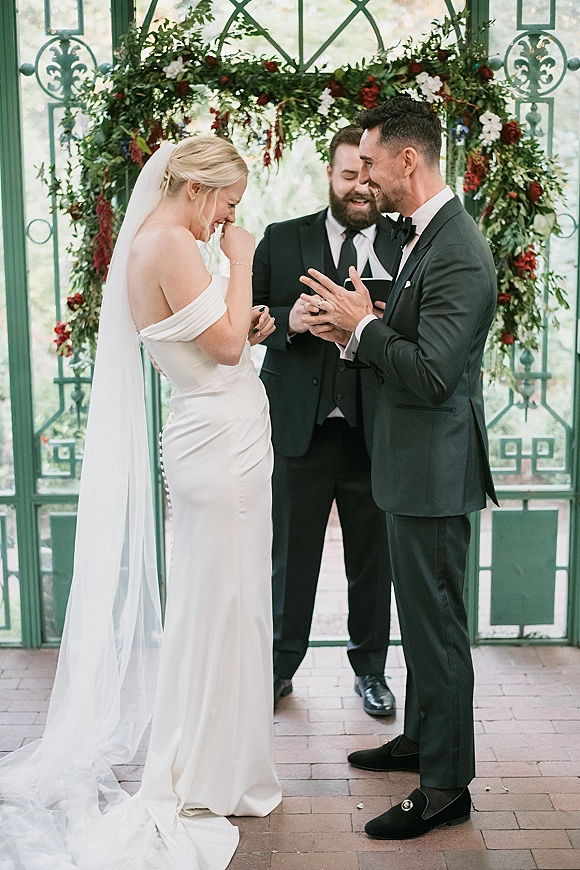 Wedding vows as bride in an off-the-shoulder gown laughs with groom in tux under a greenery arch with red flowers in a glass conservatory