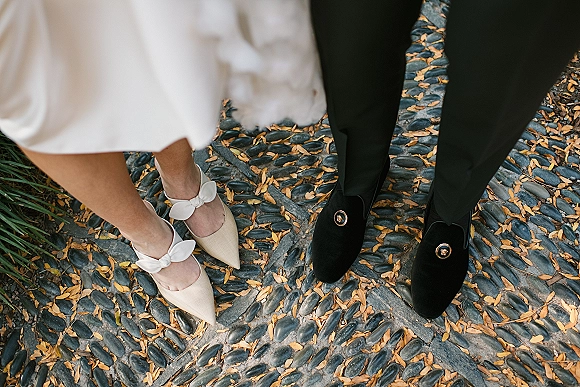 Wedding shoes beside groom loafers, ivory pointed-toe heels with bow ankle straps and black velvet crest loafers on cobblestones with fallen leaves