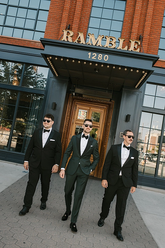 Groom and groomsmen in tuxedos and bow ties walking in sunglasses past a brick building entrance with glass windows and doors