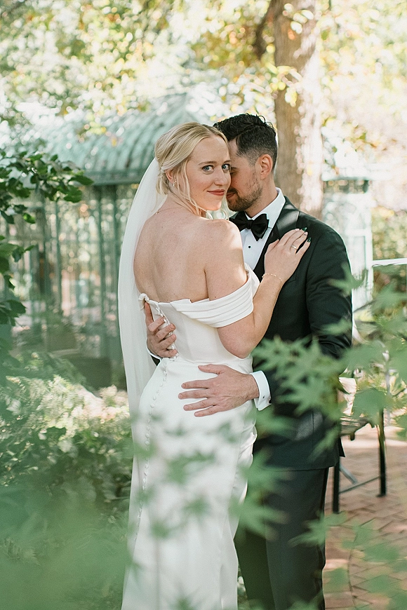 Couple portrait of bride and groom embrace as he whispers to her, veil and tuxedo in soft garden greenery by a glass greenhouse