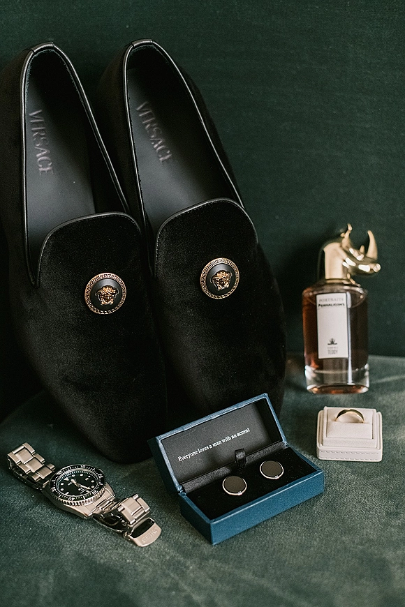 Groom accessories flat lay featuring black velvet loafers, cufflinks, wedding band, watch, and cologne on dark green fabric backdrop