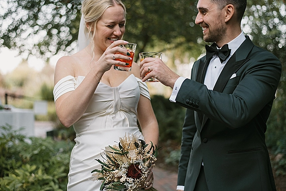 Wedding toast as bride in strapless dress and veil and groom in tuxedo clink rocks glasses on an outdoor patio with garden greenery