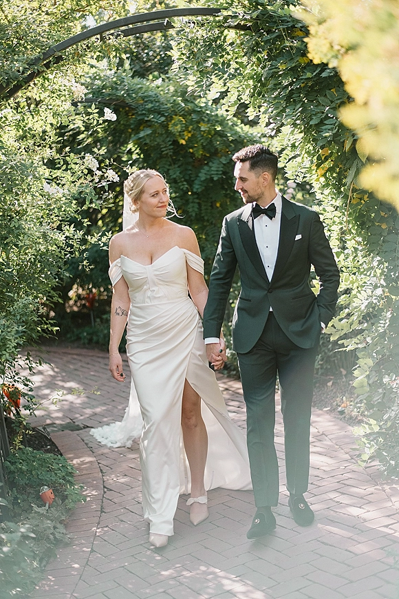Couple portrait of bride and groom walking hand in hand, veil flowing, along a sunlit brick path beneath a vine-covered arch