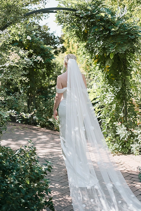 Bridal portrait of a bride from behind in an off-the-shoulder wedding dress and long veil, walking a sunlit brick path under a leafy archway