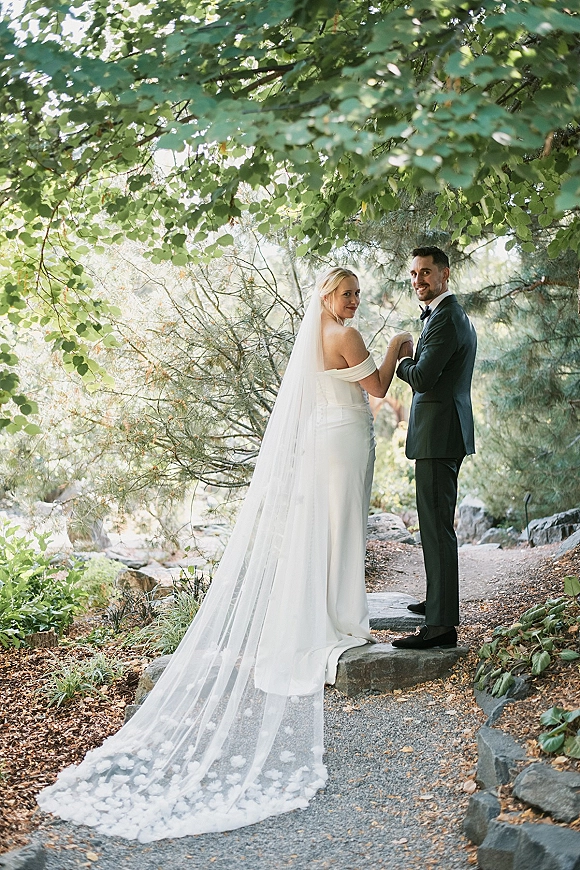 Couple portrait of bride and groom looking back, holding hands on a garden path under trees, her long veil flowing behind