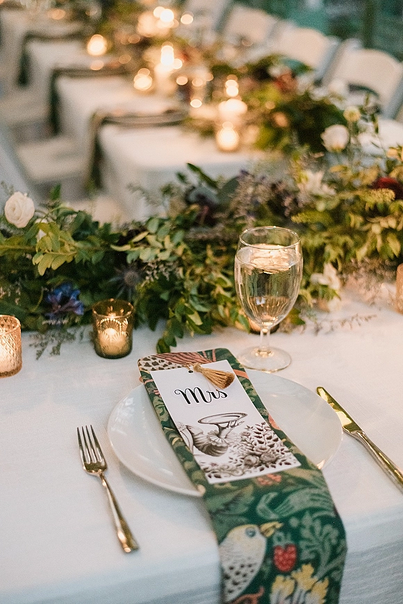 Reception tablescape with a greenery table runner, floral centerpiece and votive candles, set with white plates, gold flatware, and place cards on long banquet tables