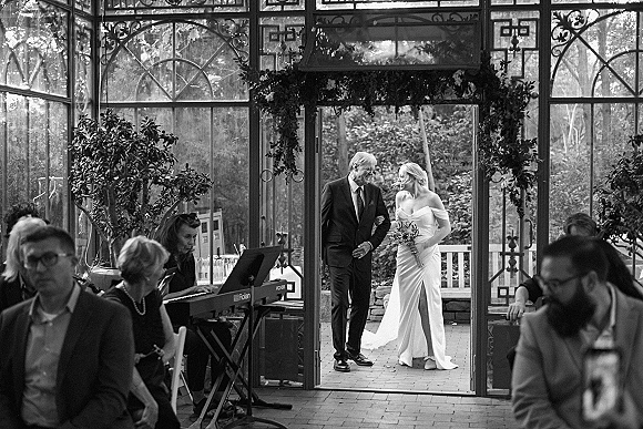 Wedding processional as bride walking down aisle with her father, holding a bouquet, entering a glass conservatory doorway past seated guests