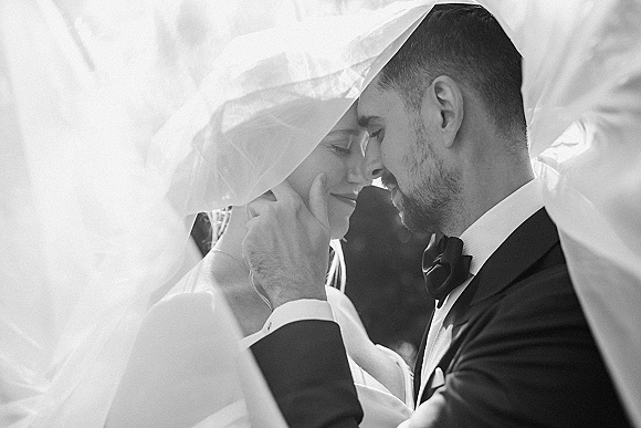 Wedding couple portrait of bride and groom close up, groom cupping her face as they touch foreheads under a veil in sunlit greenery