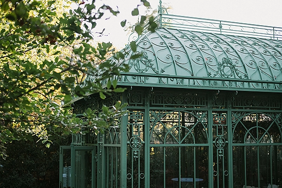 Outdoor wedding venue with a wrought iron gazebo featuring glass panels and string lights, set among sunlit garden trees and foliage