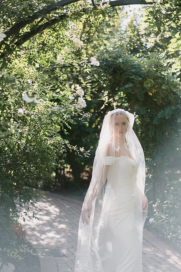 Bridal portrait of a bride with long veil in a strapless wedding dress on a sunlit brick walkway beside a garden arbor and greenery