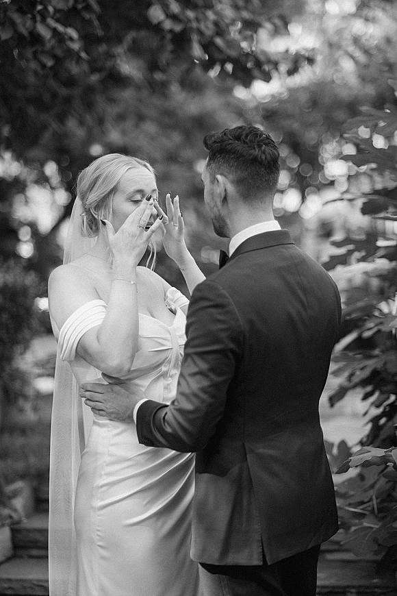 First look moment as the groom in a tuxedo comforts a crying bride wiping tears beneath her veil in an outdoor garden setting