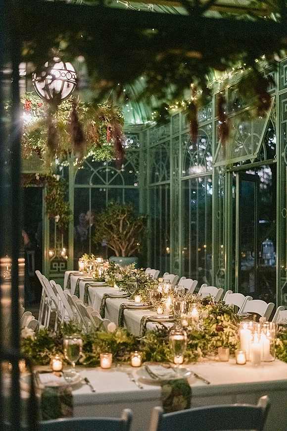 Reception tablescape with a long banquet table wedding setup, greenery garland, candles and glass hurricanes under string lights in a glass greenhouse at night