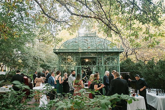 Cocktail hour with outdoor wedding cocktail hour tables in white linens beside a wedding bar setup inside a glass pavilion on a brick patio