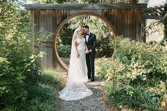 Couple portrait of bride and groom holding hands with a bridal veil as they touch foreheads by a wooden gate in garden greenery