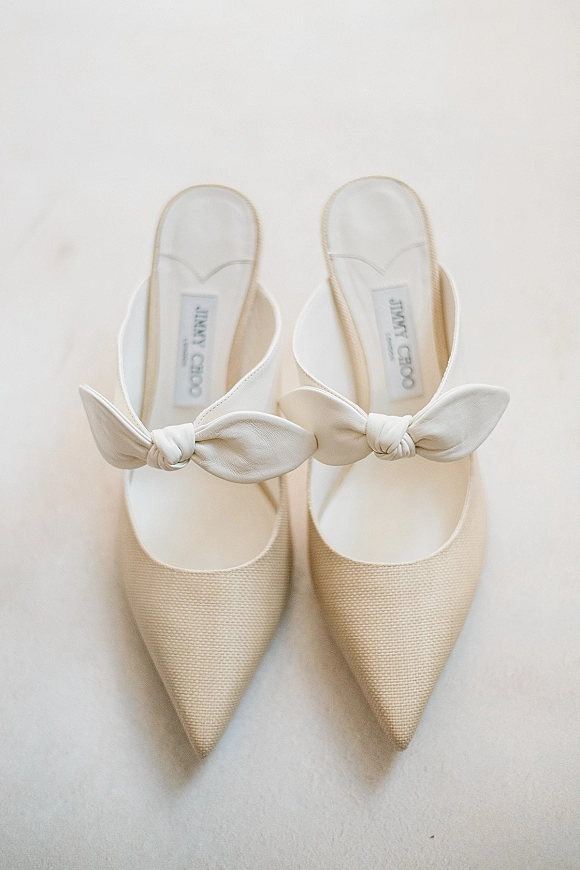 Bridal shoes with white bow heels, pointed toes and slingback straps shown close up against a clean white backdrop, fabric texture visible