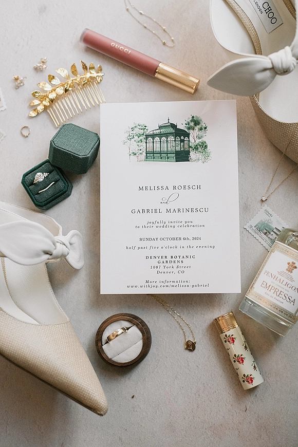 Wedding invitation suite with illustrated wedding invitation on light fabric, styled with envelope, stamp, rings, shoes, perfume and jewelry